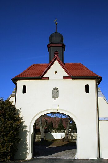 Ein Torbogen mit weißer Fassade und rotem Ziegeldach. Auf dem Dach ein kleines schwarzes Türmchen.