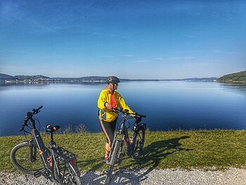 Eine Frau steht neben ihrem Fahrrad am Ufer eines Sees. Neben ihr steht ein weiteres Fahrrad. Hinter ihr ein blauer See unter blauem Himmel.