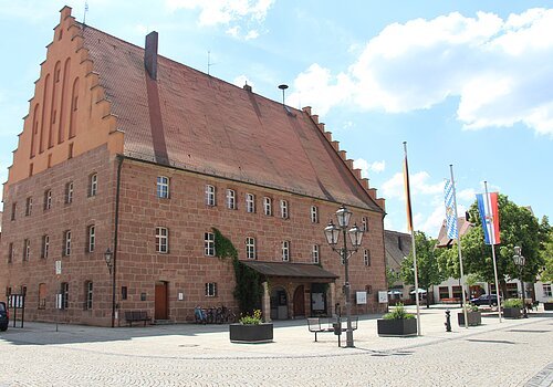 Großes historisches Backsteingebäude mit Treppengiebel und drei Flaggen auf einem gepflasterten Platz bei Tageslicht.