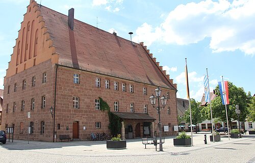 Großes historisches Backsteingebäude mit Treppengiebel und drei Flaggen auf einem gepflasterten Platz bei Tageslicht.